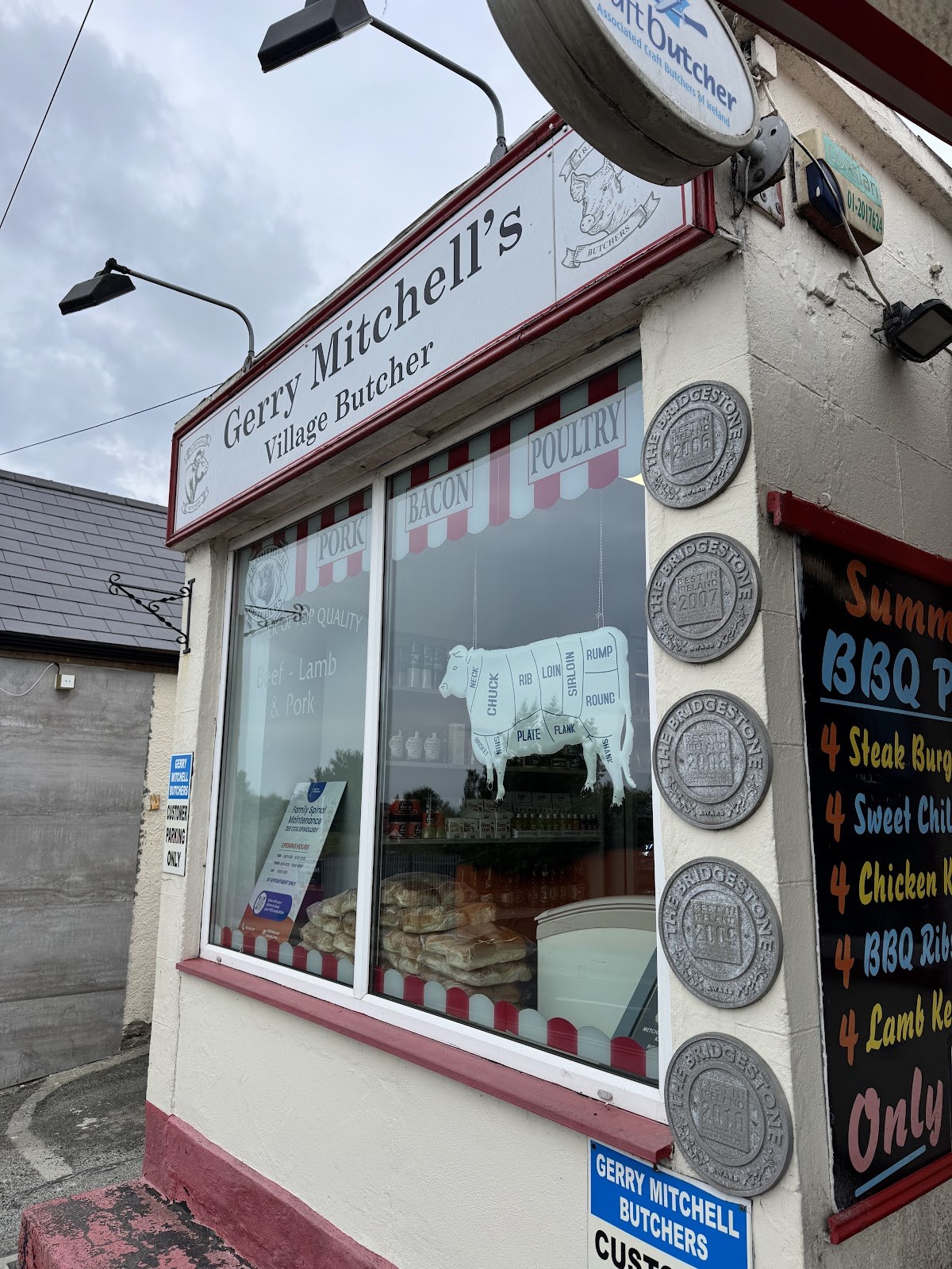 Shopfront of Gerry Mitchell’s Village Butcher on Main Street, Kilcoole, with a beef-cut diagram in the window and a Summer BBQ board